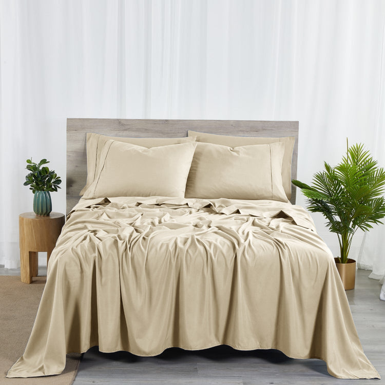 A warm taupe colored 6-piece bamboo sheet set on a bed, with a flat sheet, fitted sheet, and pillowcases displayed. The bed is set against a room backdrop with a plant and nightstand on the side.