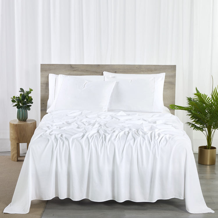 A white 6-piece bamboo sheet set on a bed, including a fitted sheet, flat sheet, and pillowcases, presented in a bright room setting with decorative plants and a nightstand.