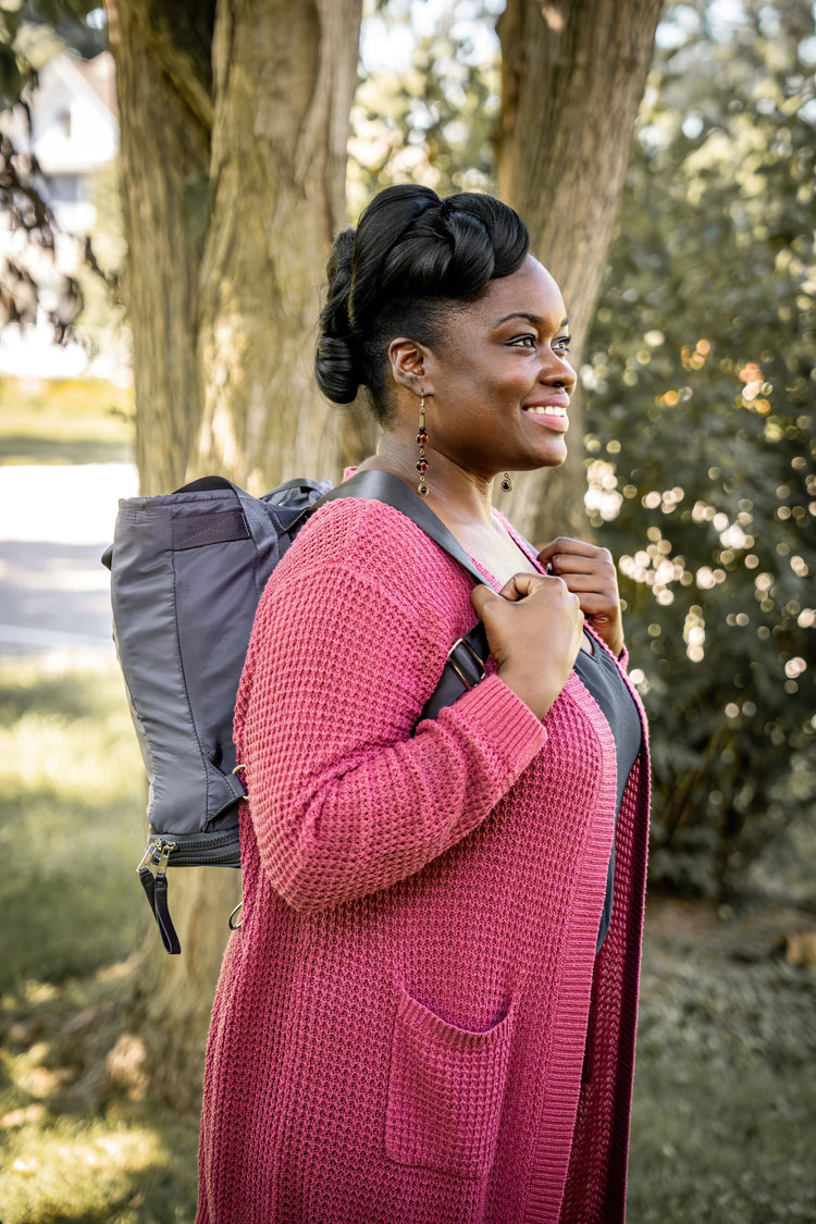 Woman in a pink cardigan with a gray backpack standing outdoors near trees.