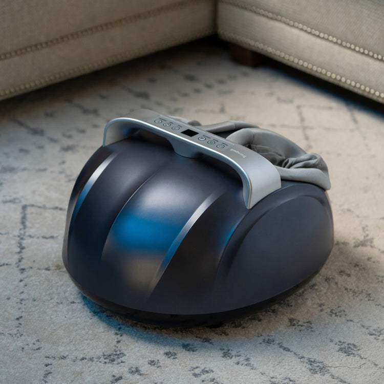 A dark-colored foot massager with a smooth, ergonomic design, featuring a footrest and control panel, placed on a light-colored carpet.