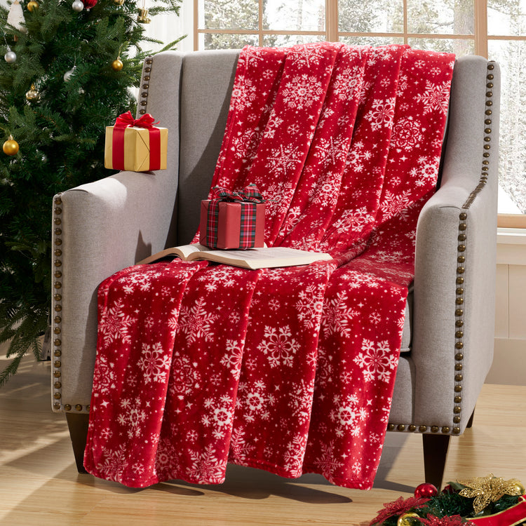 Red blanket with white snowflake pattern draped over a gray armchair next to a Christmas tree and presents.