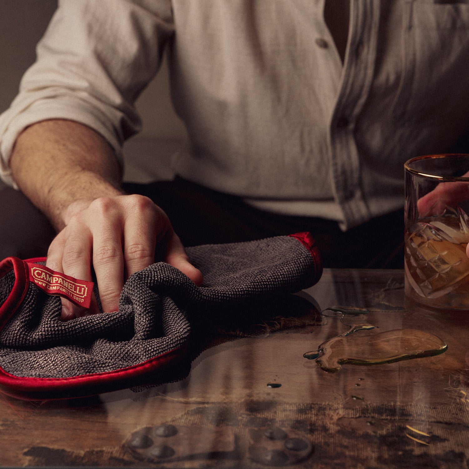 Person cleaning a glass with a Campari branded towel on a wooden table.