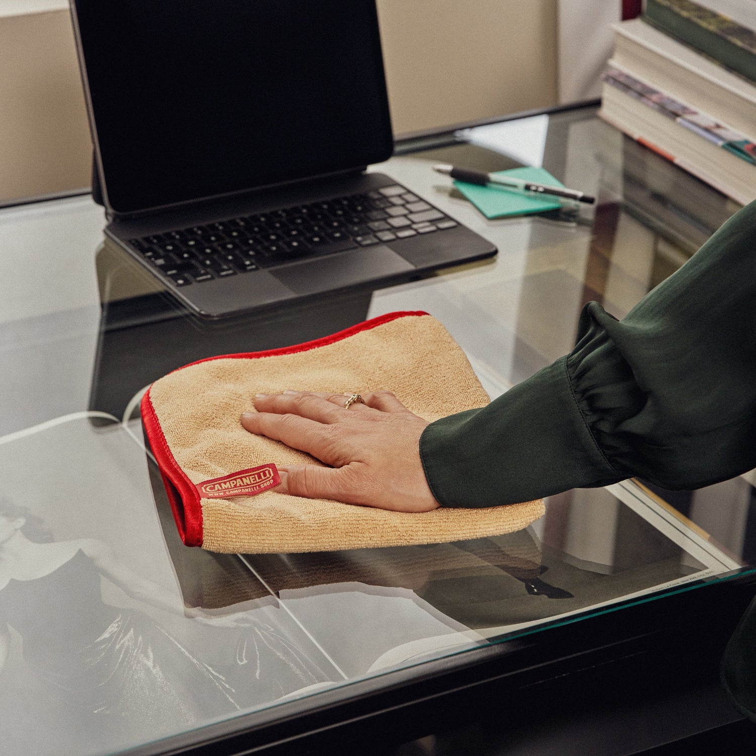 Person cleaning a glass desk with a microfiber cloth, laptop and books in the background.