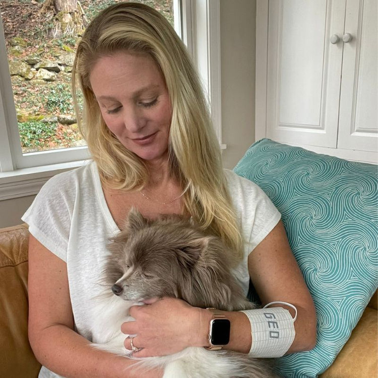Woman holding a small dog in a home setting with a window and blue pillow in the background.