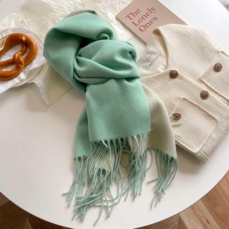 A green reversible pashmina scarf with fringe detailing, displayed on a white surface next to a beige coat and a pretzel.