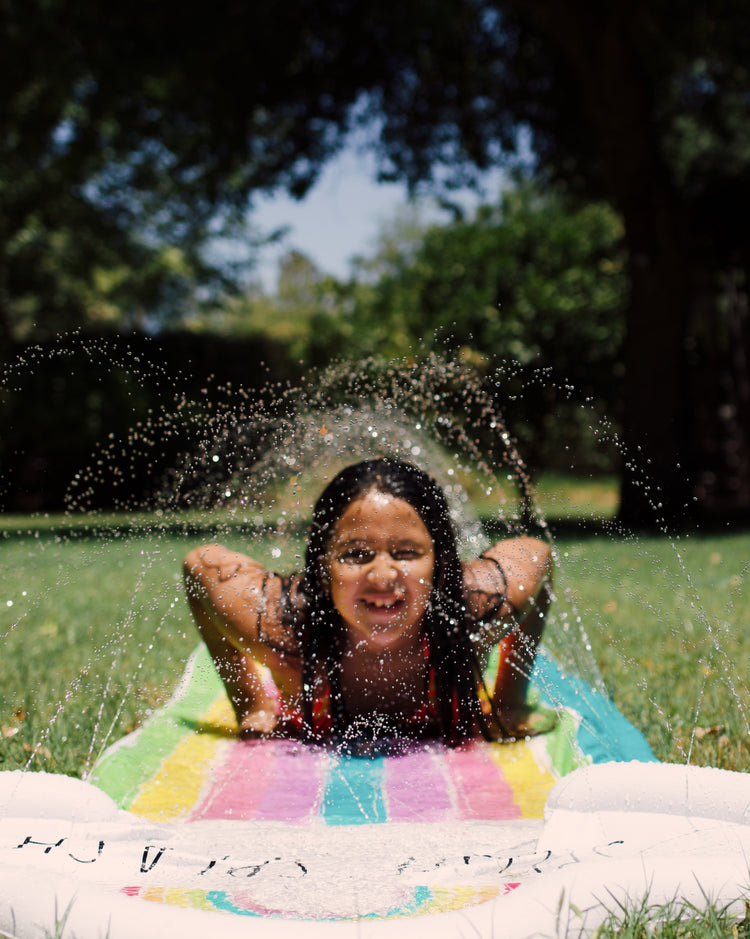 Child playing in a sprinkler on a colorful inflatable pool toy in a park.