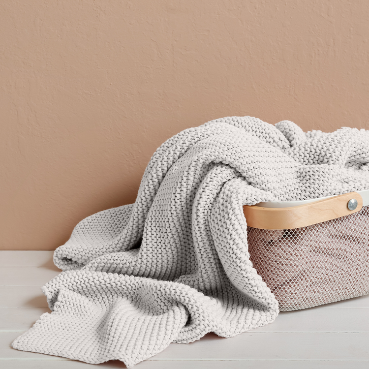 Gray knitted blanket draped over a woven basket on a beige surface with a beige wall background