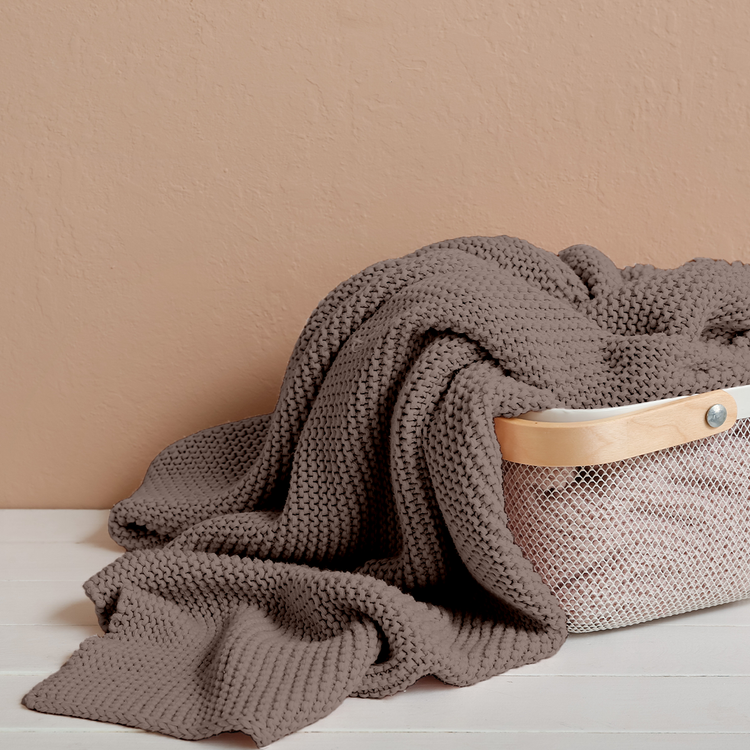 Brown knitted blanket draped over a woven basket on a beige surface.