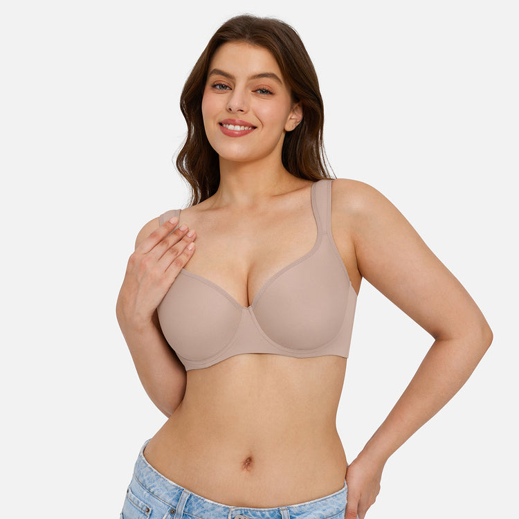 A woman modeling a seamless, wireless, scoop-neck bra in a light taupe-beige color, paired with light-wash blue jeans against a solid light gray background.