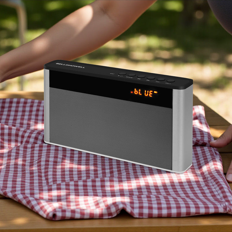 Black portable speaker on a red and white checkered cloth with a blurred outdoor background