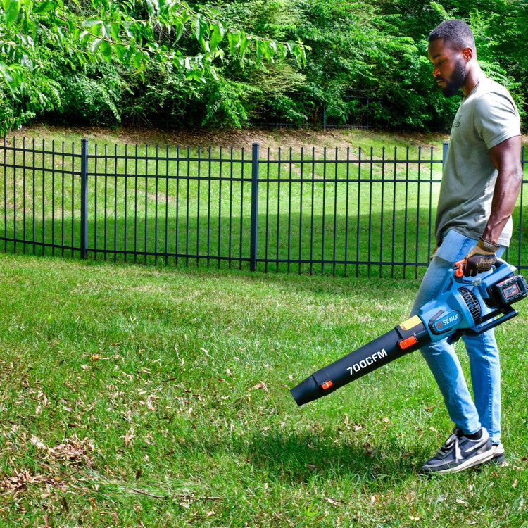 Man using a leaf blower in a grassy area with a fence and trees in the background