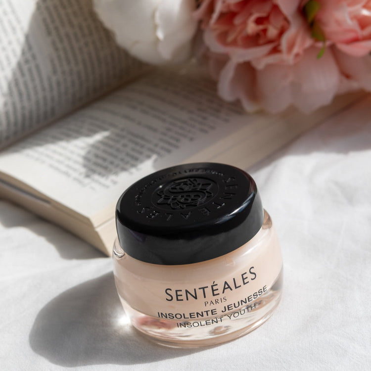 Cosmetic jar with black lid on a white surface with flowers and an open book in the background