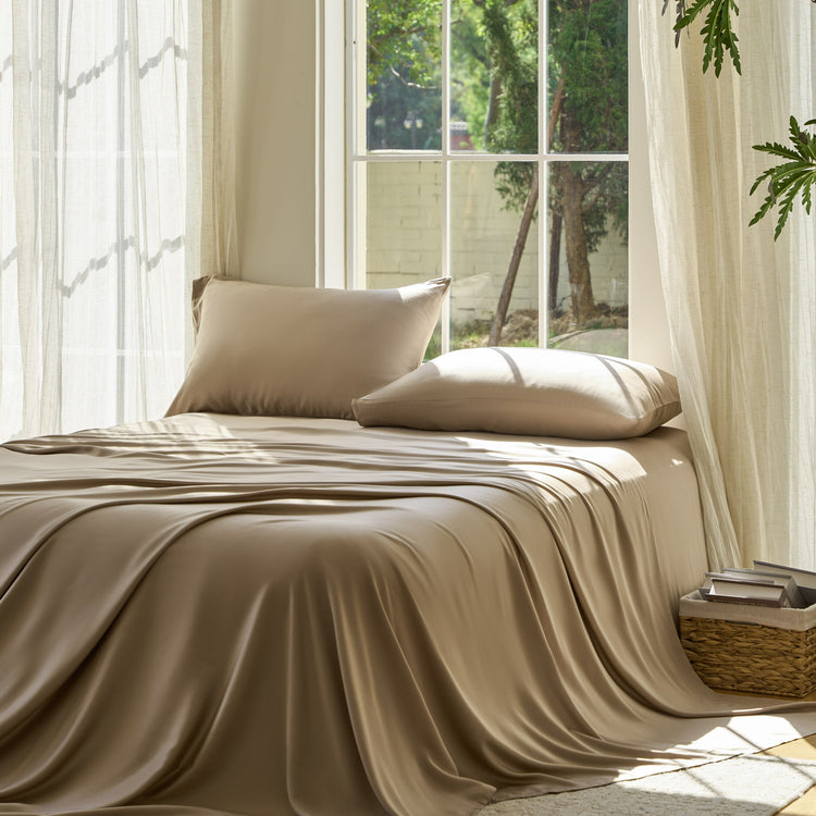Beige bedding set on a bed with pillows in a bright room with large windows.