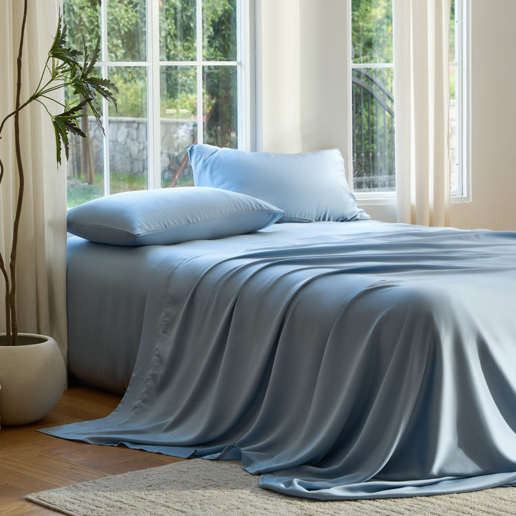 Bed with light blue bedding and pillows in a bright room with large windows.