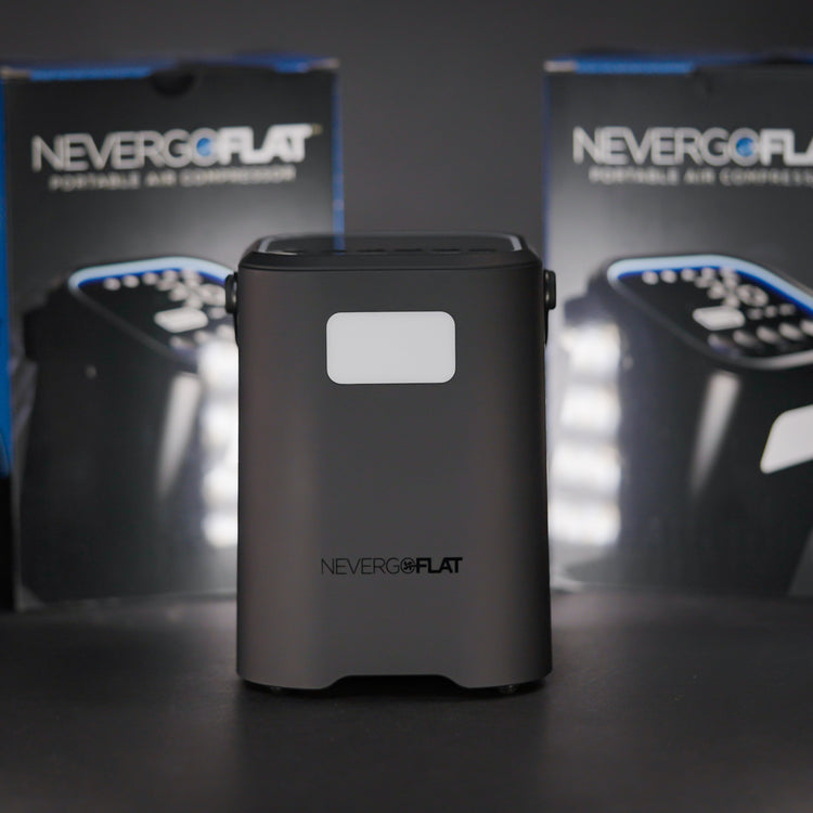 A portable air compressor branded NEVERGOFLAT, with packaging boxes in the background. The device is black, featuring a digital display, and appears to be compact and sturdy.