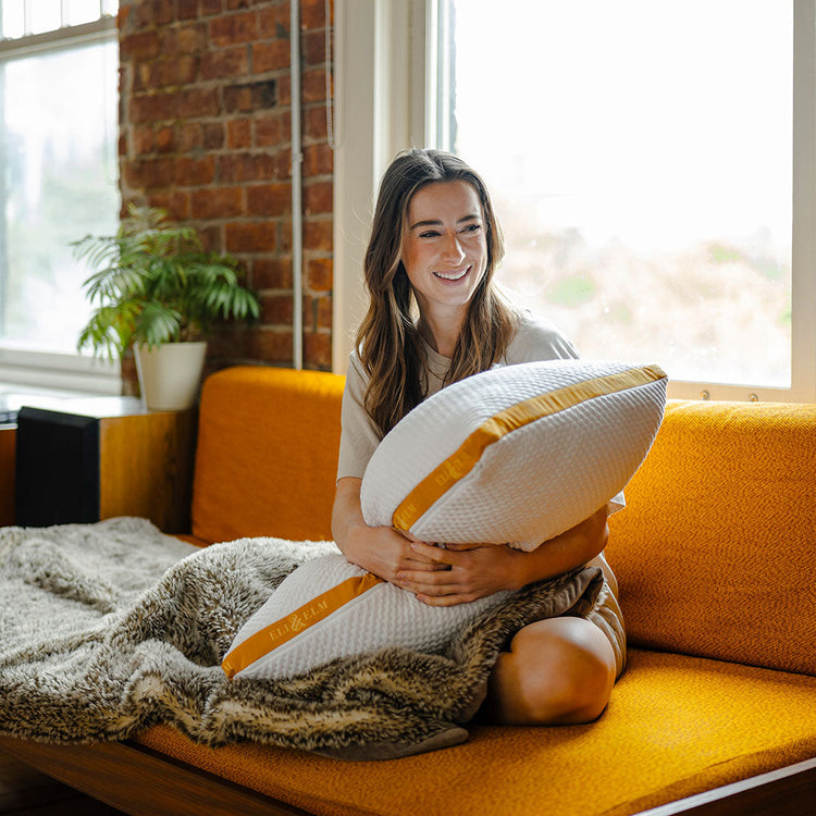 A woman sitting on a yellow couch, hugging a white pillow with a geometric pattern.