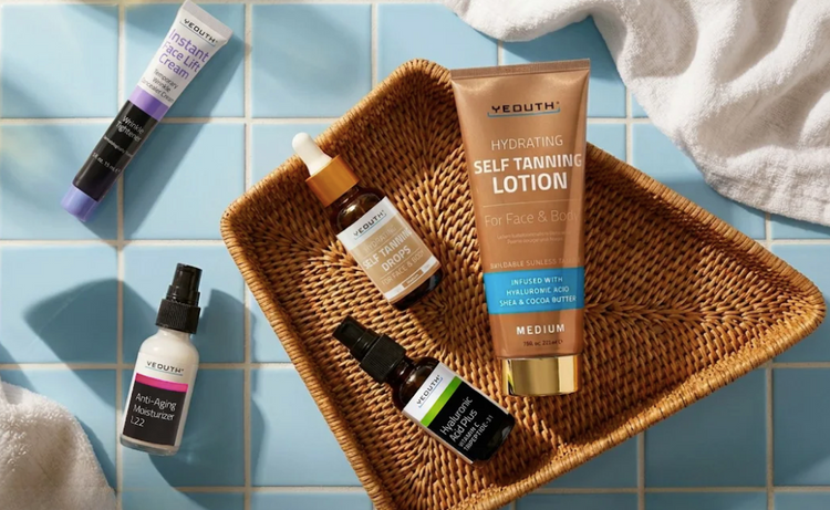 Collection of self-tanning products including a bottle of Vudhu Hydrating Self-Tanning Lotion on a woven tray with a blue tiled background.