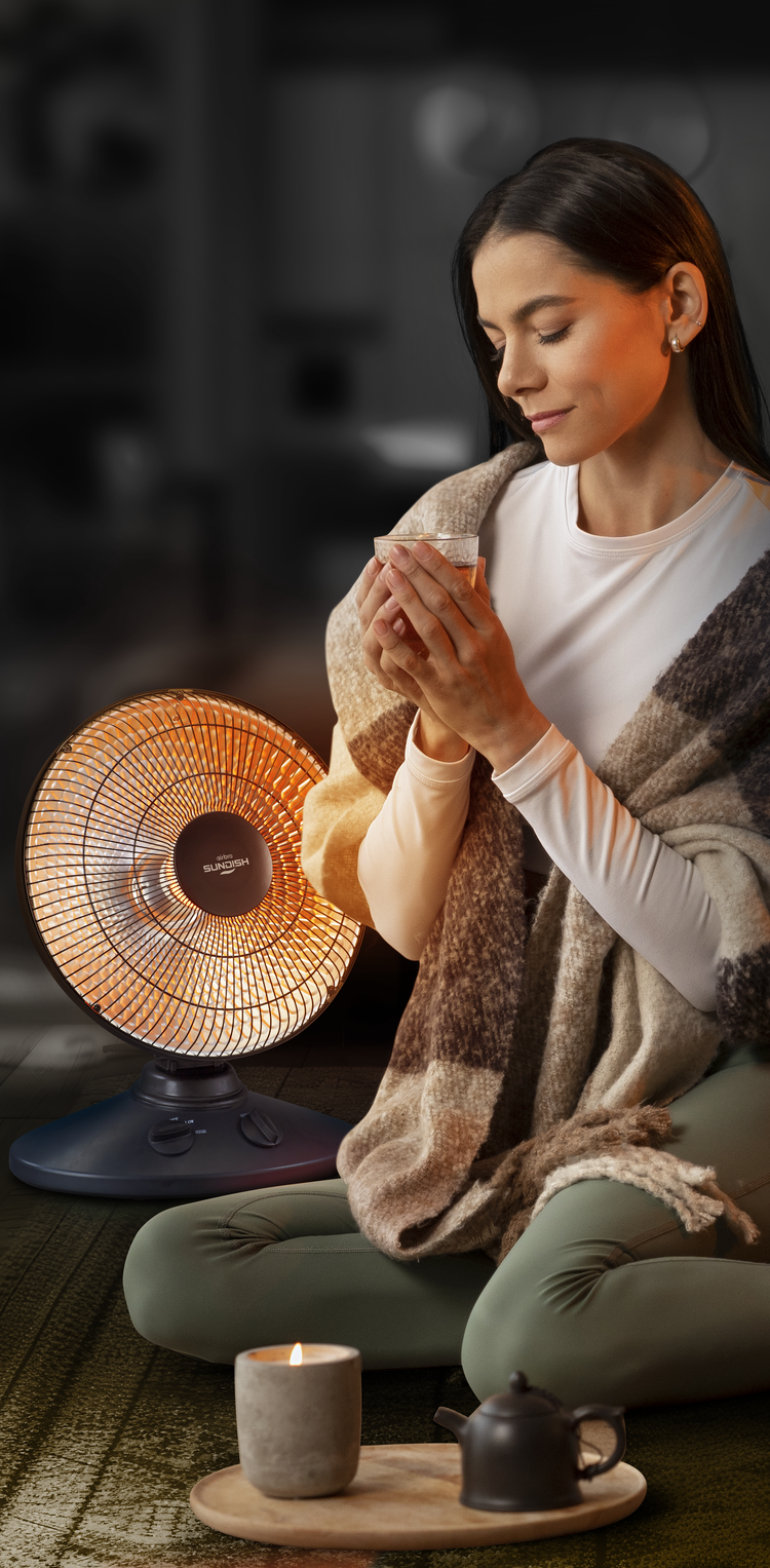 Woman sitting on the floor with a small heater, cup, and tea set in a cozy setting.
