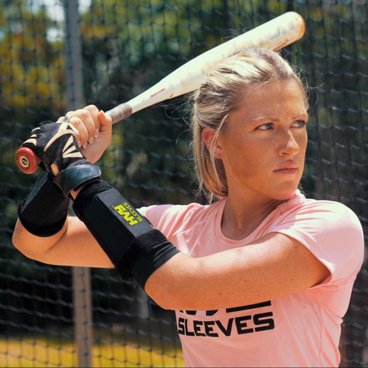 Woman holding a baseball bat with a black arm sleeve, standing in a batting cage.