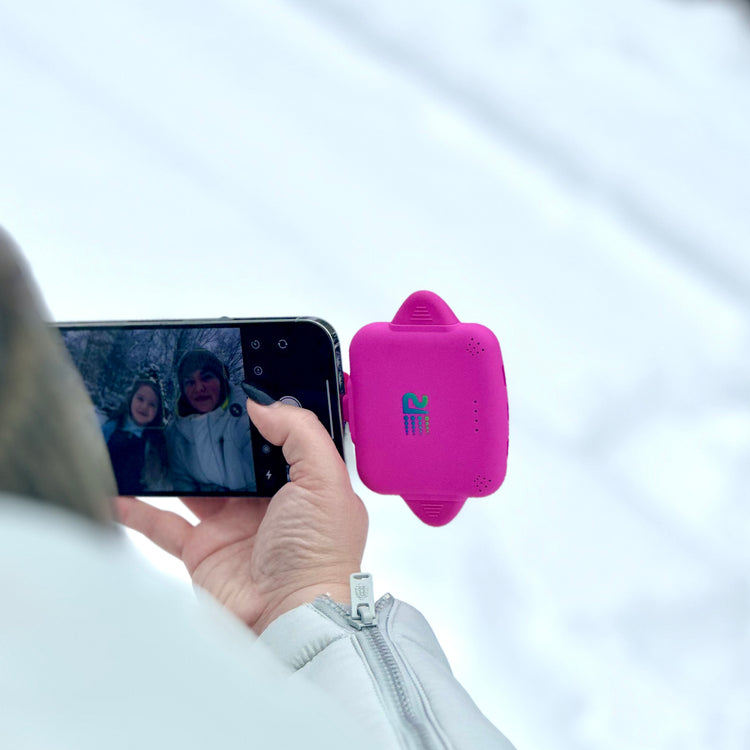 A person outdoors in a snowy setting holds a smartphone showing a video call while wearing a bright pink smartwatch with a small blue and green logo on the front.
