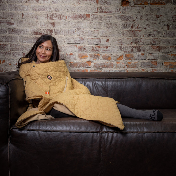Woman sitting on a couch with a yellow quilt draped over her, against a brick wall.