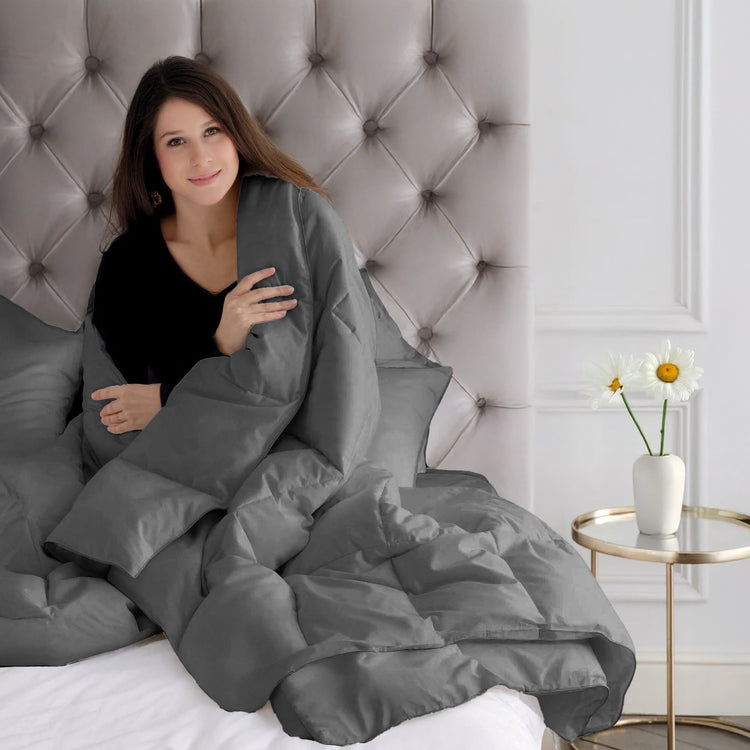 Woman wrapped in a gray comforter sitting on a tufted headboard in a bedroom.