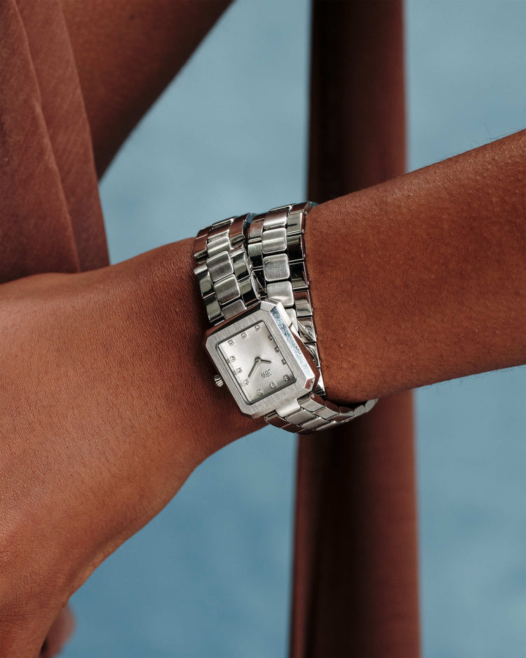A close-up shows a persons wrist with medium skin tone wearing the JBW Watches Arc Double | J6390J, a square-faced silver watch with a metal band, set against a soft blue background and partially draped in brown fabric.