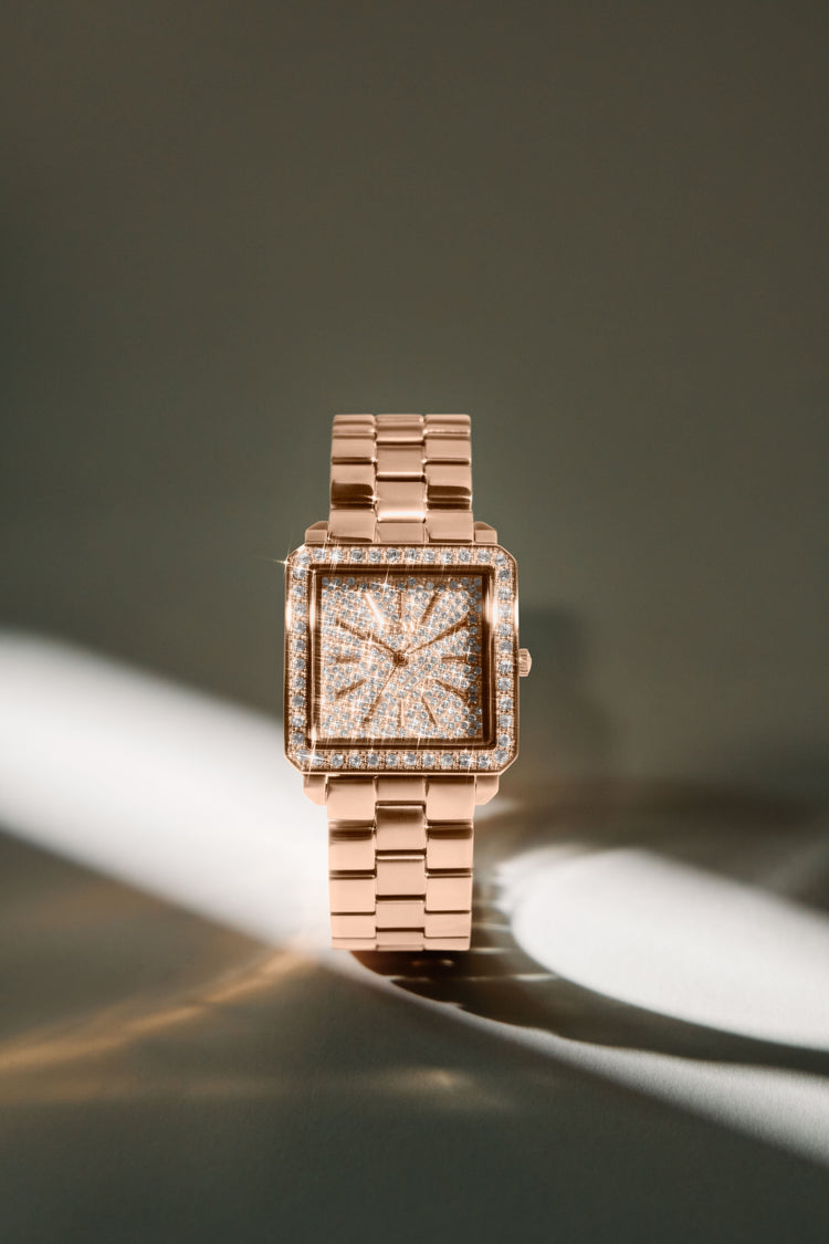 A square rose gold watch with a diamond-encrusted face and matching rose gold link bracelet, photographed on a dark gray background with dramatic diagonal light beams.