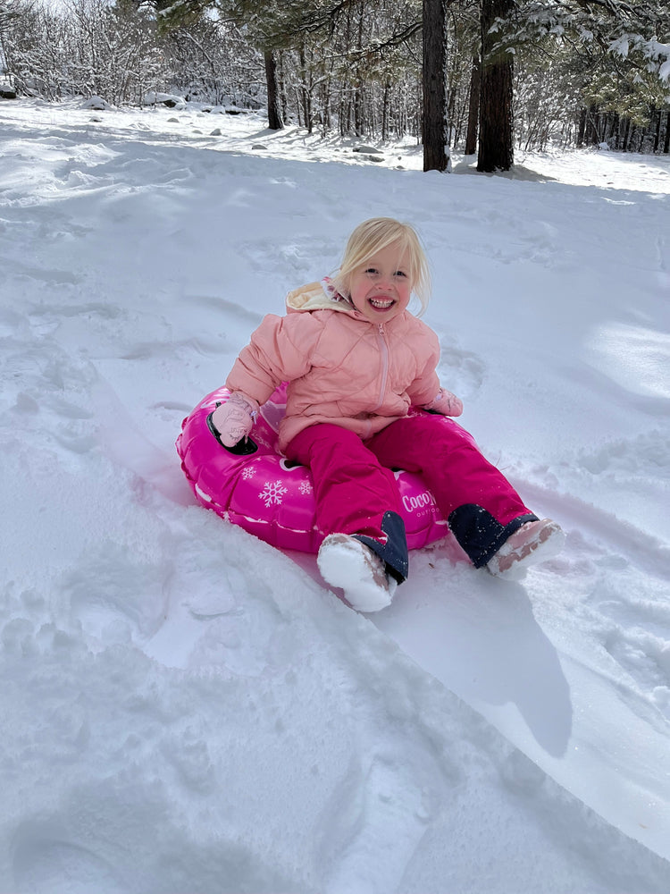 Child in pink snowsuit sitting on a sled in a snowy forest