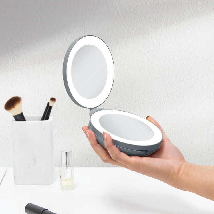 Hand holding a round, illuminated makeup mirror with a compact design on a neutral background.