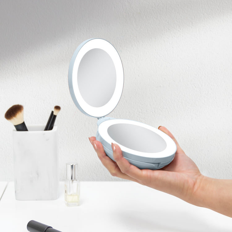 Hand holding a round, illuminated makeup mirror with a light gray base on a white background.