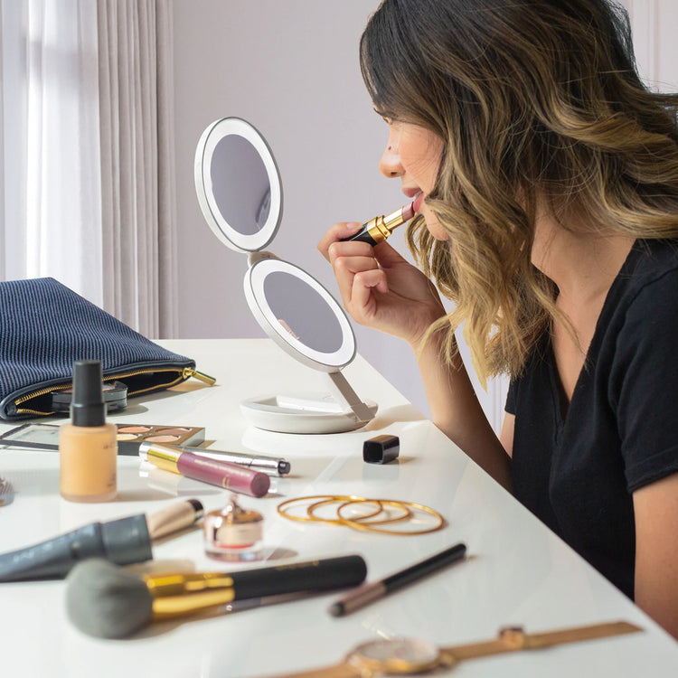 Woman applying lipstick in front of a makeup mirror with various products on a table.