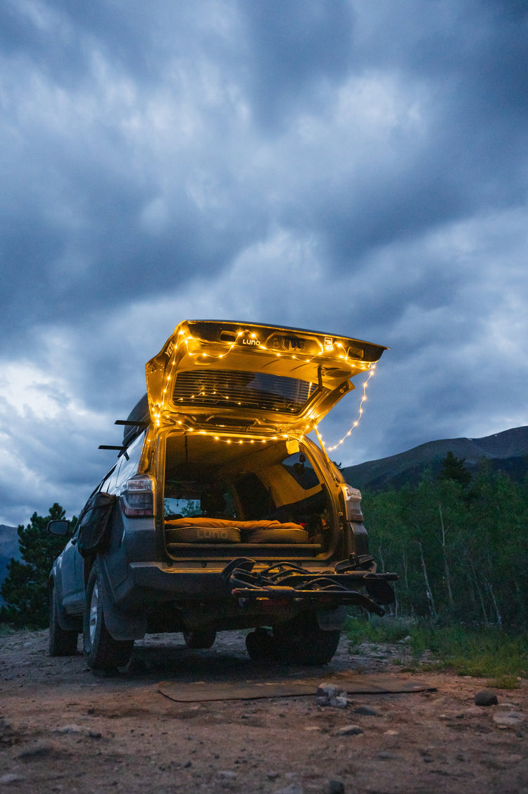 Car with open trunk illuminated by string lights against a mountainous landscape under a cloudy sky.