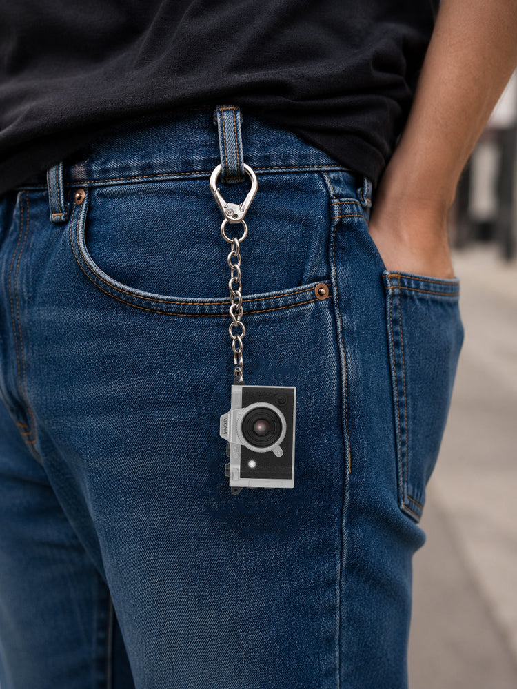 A silver metal camera-shaped keychain with a black lens detail hangs from a silver ring attached to the back pocket of blue jeans, shown on a person wearing a black t-shirt.