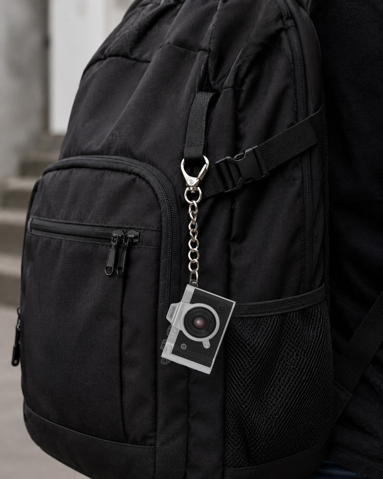 A close-up of a black backpack with a silver camera-shaped keychain attached to a strap, shown against a blurred outdoor background.