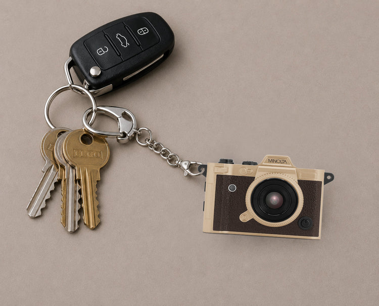 A novelty keychain featuring a miniature vintage camera charm attached to a silver split ring with a car key fob and two keys, displayed on a neutral beige background.
