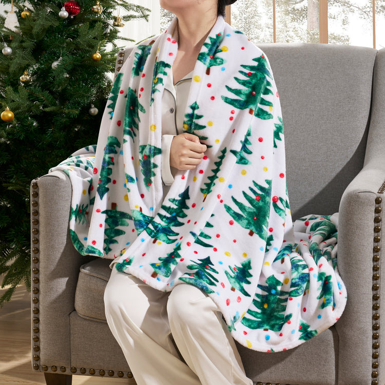 Person sitting on a couch with a Christmas-themed blanket draped over them, next to a decorated Christmas tree.