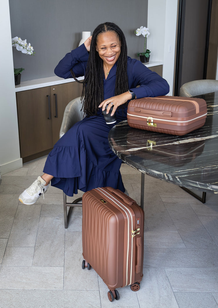 Woman in a blue dress sitting at a table with two brown suitcases in a room.