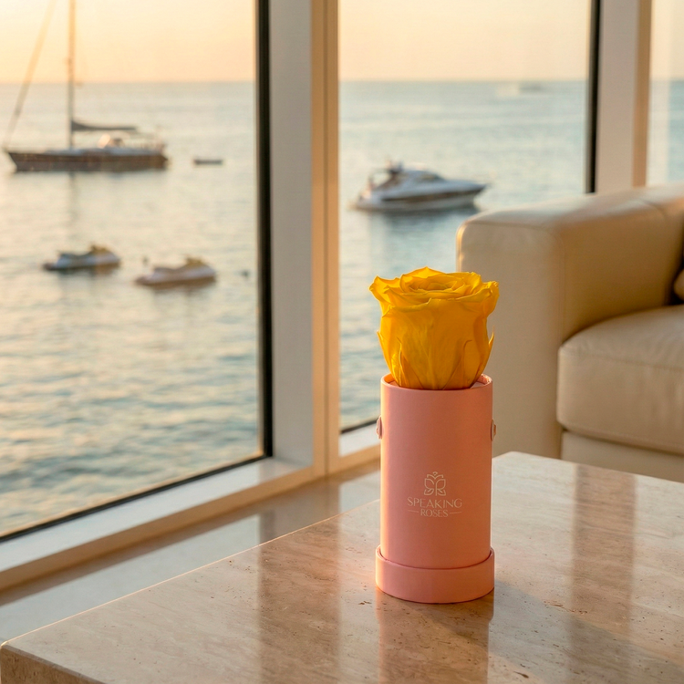 Pink vase with yellow flowers on a table by a large window with a view of the sea and boats.