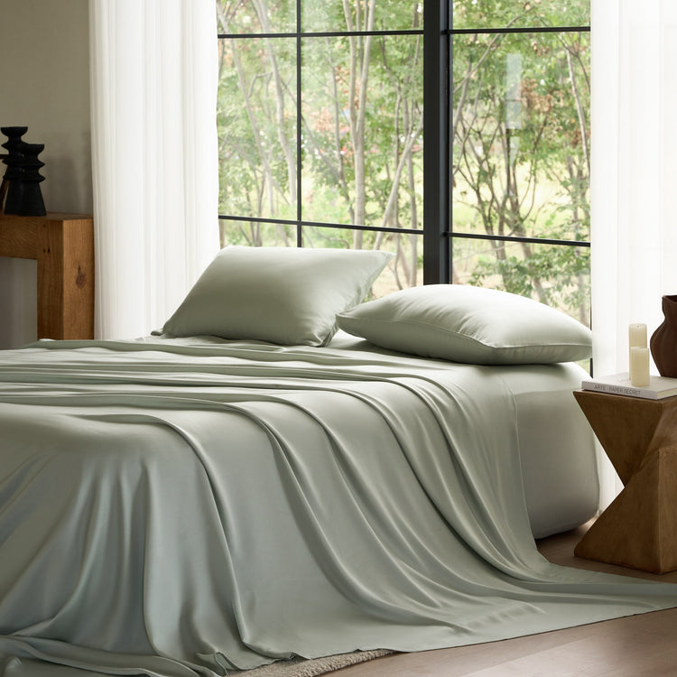 Bed with light green bedding and pillows in a room with large windows showing trees.