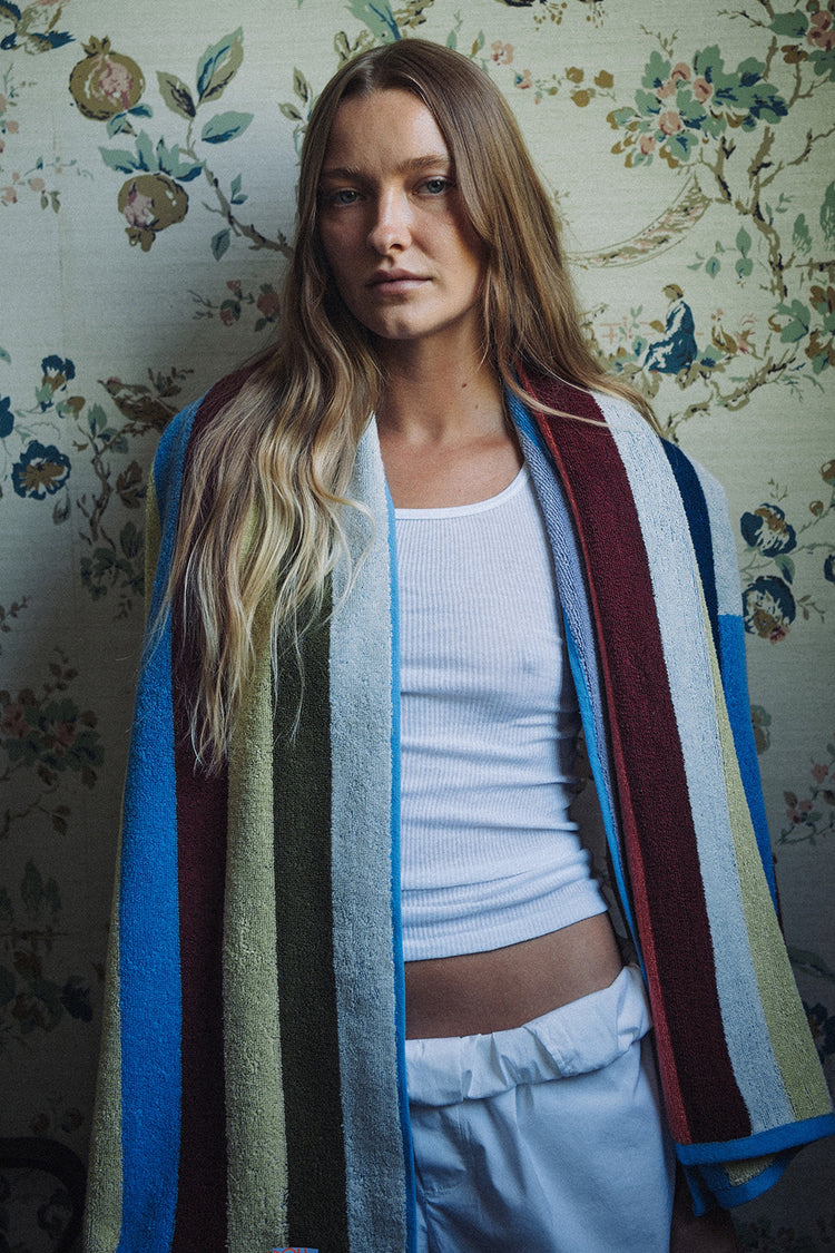 Woman wearing a colorful striped cardigan over a white top and light blue pants against a floral wallpaper background.
