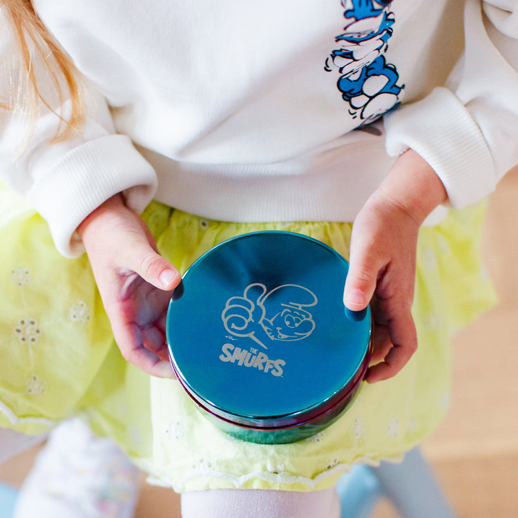 Child holding a blue container with 'Smurfs' logo, wearing a white sweater with Smurfs.