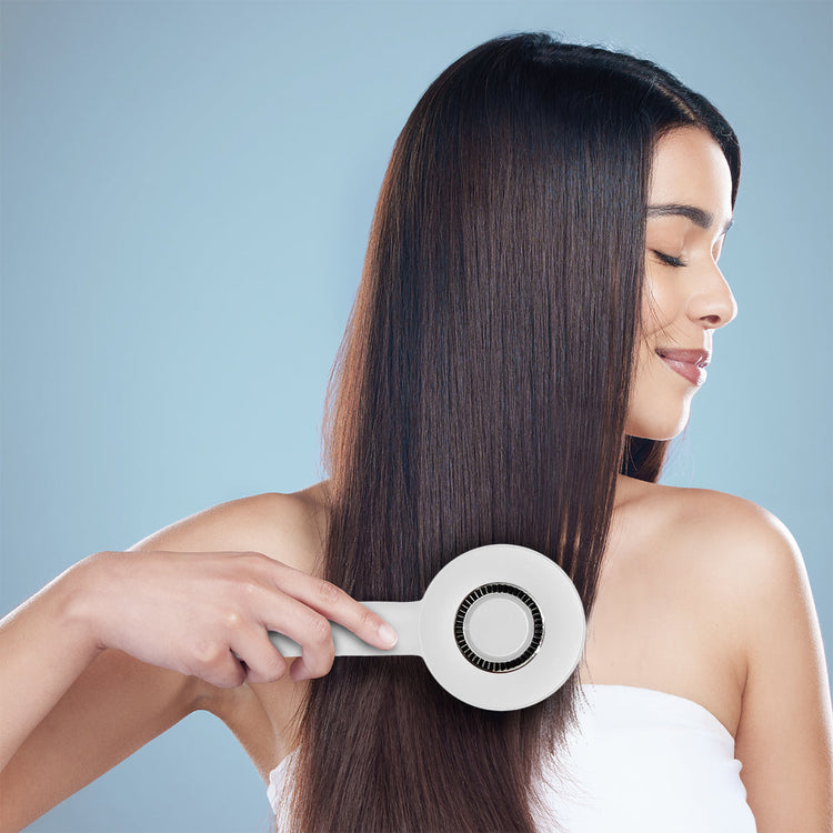 Woman using a hair straightening brush on her long, straight hair against a light blue background