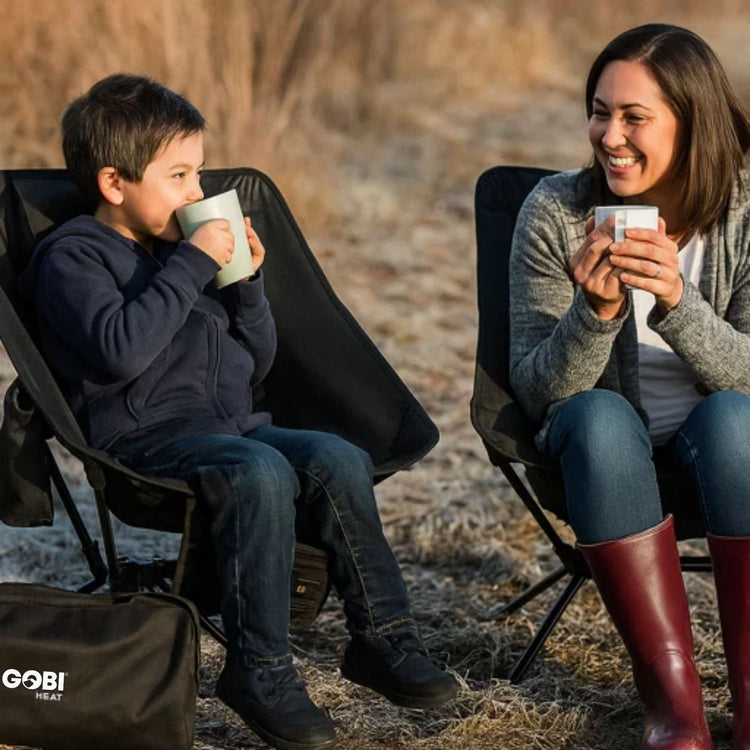 Two people sitting in camping chairs outdoors, drinking from mugs.