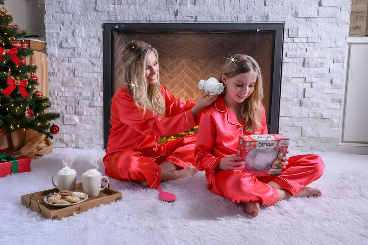 Two women in red pajamas sitting on a white rug in front of a fireplace, with a Christmas tree in the background.