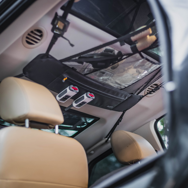 The interior of an adventure mobile showcasing the Luno® Cargo Hammock attached to the ceiling above the rear seats. The hammock, perfect for gear storage, is black with various compartments and two labeled white straps with red clips. The car seats are tan in color.