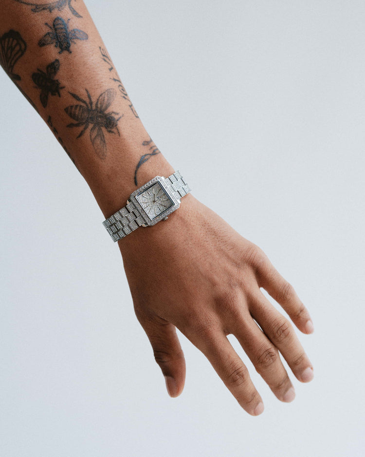 A tattooed arm with an extended hand wears the JBW Cristal 28 | J6386C, a sleek stainless steel wristwatch with a rectangular face and metal band, set against a plain light gray background.