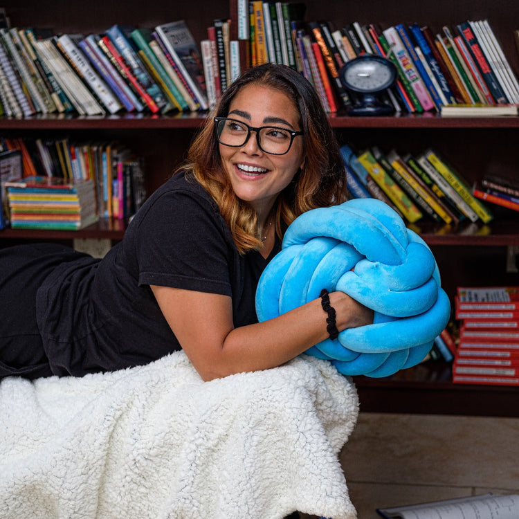 Woman holding a blue knot-shaped pillow in front of a bookshelf