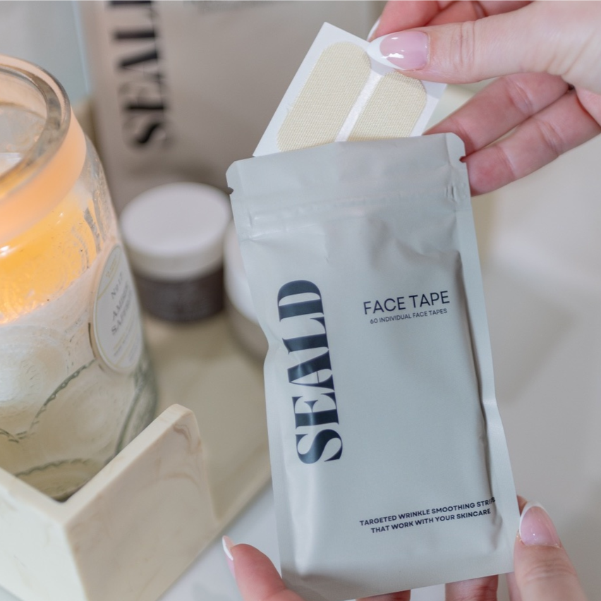 Hand holding a package of 'SEALD' face tape with a candle in the background
