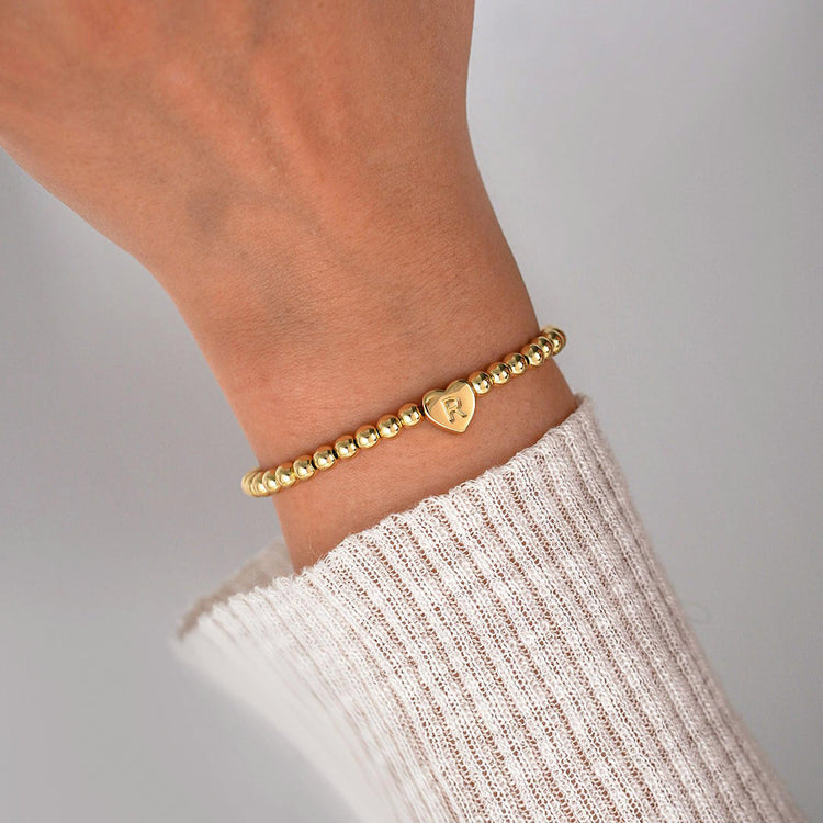 Gold bracelet with a heart-shaped charm on a wrist against a neutral background
